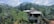 A house is situated amidst lush greenery with dense trees and palm trees around. The structure has a sloped roof with red shingles and appears to be partially constructed or undergoing renovation. In the background, a hill rises under a partly cloudy sky.