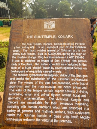 A large, weathered informational plaque situated in a lush, green outdoor setting. The text on the plaque describes the historical significance of the Sun Temple in Konark, Odisha, noting its construction in the 13th century during the reign of King Narasimhadeva. It highlights the temple's architectural features, including carvings and sculptures, and its representation of the Sun-god.