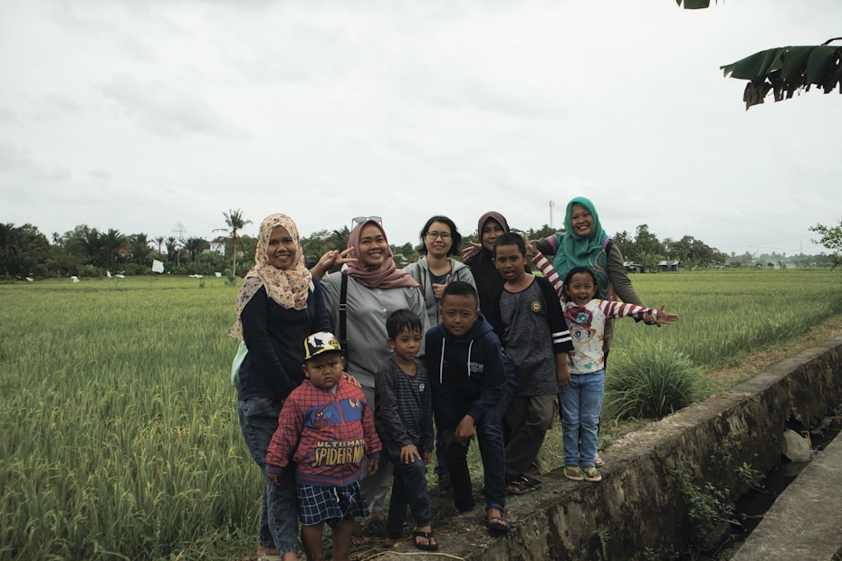 A group of people, including adults and children, poses happily in a lush green field, with long grass and a cloudy sky. The adults wear casual clothing and headscarves, while the children wear colorful shirts and jackets. The scene captures a sense of unity and togetherness in a natural setting.