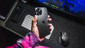 A hand holds a sleek black smartphone with a triple camera setup. The phone has a distinctive logo on its back. The background features a desk setup including a desktop keyboard, a wireless mouse, a monitor displaying a graphic, and a phone case. The person is wearing a colorful, tie-dyed sweatshirt.
