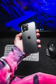 A smartphone with a glossy, black finish is being held in a human hand, displaying the iconic Apple logo on the back. The person wearing a vibrant tie-dye sweater is positioned in front of a backlit keyboard and mouse. In the background, a digital graphic featuring abstract blue shapes and lines is visible, possibly a computer screen.