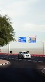 A white car is driving on a curved road near a roundabout. Overhead, there are blue and white directional signs indicating routes to Old Airport and Al Hilal - Najma, as well as a sign for Doha International Airport. Trees and landscaping are present on the left, while a security fence runs in the background.