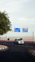 A white car is driving on a curved road near a roundabout. Overhead, there are blue and white directional signs indicating routes to Old Airport and Al Hilal - Najma, as well as a sign for Doha International Airport. Trees and landscaping are present on the left, while a security fence runs in the background.