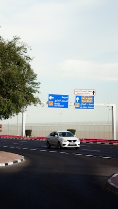 A white car is driving on a curved road near a roundabout. Overhead, there are blue and white directional signs indicating routes to Old Airport and Al Hilal - Najma, as well as a sign for Doha International Airport. Trees and landscaping are present on the left, while a security fence runs in the background.