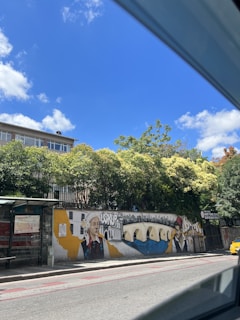 A vibrant mural is painted on a roadside wall featuring traditional and modern elements. The artwork includes portraits of two individuals in cultural attire, a river with a bridge, and a cityscape in the background. Trees provide a lush backdrop, and a clear blue sky with scattered clouds is visible above.