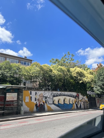 A vibrant mural is painted on a roadside wall featuring traditional and modern elements. The artwork includes portraits of two individuals in cultural attire, a river with a bridge, and a cityscape in the background. Trees provide a lush backdrop, and a clear blue sky with scattered clouds is visible above.