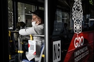 A person wearing a white shirt and black cap, masked, is seated inside a bus, gazing out from the side window. The bus features a large G20 logo with 'Indonesia 2022' written below it. A QR code is attached to the back seat.