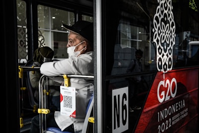 A person wearing a white shirt and black cap, masked, is seated inside a bus, gazing out from the side window. The bus features a large G20 logo with 'Indonesia 2022' written below it. A QR code is attached to the back seat.