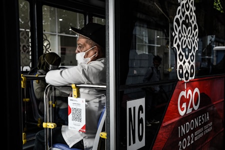 A person wearing a white shirt and black cap, masked, is seated inside a bus, gazing out from the side window. The bus features a large G20 logo with 'Indonesia 2022' written below it. A QR code is attached to the back seat.