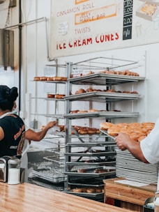 A bakery setting with two people, one wearing a black t-shirt and the other in a white shirt. They are handling trays filled with assorted donuts, stacked on metal racks. A menu on the wall offers items like cinnamon rolls and ice cream cones. The countertop in the foreground has a container of sugar.