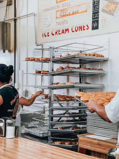 A bakery setting with two people, one wearing a black t-shirt and the other in a white shirt. They are handling trays filled with assorted donuts, stacked on metal racks. A menu on the wall offers items like cinnamon rolls and ice cream cones. The countertop in the foreground has a container of sugar.