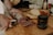 A person with tattooed hands is slicing a piece of cured sausage on a wooden cutting board. The sausage has a marbled appearance. In the background, there are slices of bread, a jar with a dark liquid, a bottle of olive oil, and various spices.