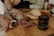 A person with tattooed hands is slicing a piece of cured sausage on a wooden cutting board. The sausage has a marbled appearance. In the background, there are slices of bread, a jar with a dark liquid, a bottle of olive oil, and various spices.