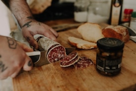 A person with tattooed hands is slicing a piece of cured sausage on a wooden cutting board. The sausage has a marbled appearance. In the background, there are slices of bread, a jar with a dark liquid, a bottle of olive oil, and various spices.