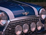 A close-up of a classic car's front, exhibiting a glossy blue paint with white racing stripes. There are four circular headlights and a chrome grille that enhance the vintage aesthetic. The manufacturer's emblem, which features wings and a red circle, is prominently displayed.
