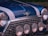 A close-up of a classic car's front, exhibiting a glossy blue paint with white racing stripes. There are four circular headlights and a chrome grille that enhance the vintage aesthetic. The manufacturer's emblem, which features wings and a red circle, is prominently displayed.