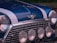 A close-up of a classic car's front, exhibiting a glossy blue paint with white racing stripes. There are four circular headlights and a chrome grille that enhance the vintage aesthetic. The manufacturer's emblem, which features wings and a red circle, is prominently displayed.