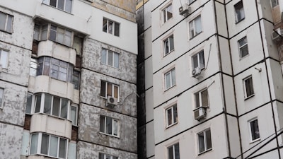 Two adjacent apartment buildings are shown. The building on the left has a weathered and worn appearance, with peeling paint and an assortment of variously shaped and sized windows. The building on the right appears more modern, with cleaner surfaces and uniform windows. Both buildings have air conditioning units mounted on the exterior.