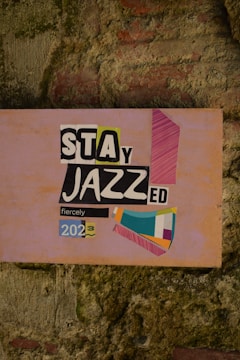 A wall with a textured, rustic appearance featuring a colorful poster that reads 'Stay Jazzed fiercely 2023'. The poster combines various fonts and colors, including black, white, yellow, and pink elements on a pinkish background, along with abstract geometric shapes.