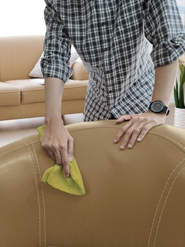 A person is seen cleaning a beige leather sofa with a yellow cloth. The individual is wearing a plaid shirt and a watch. The background includes a beige couch with cushions and a plant, indicating a living room setting.