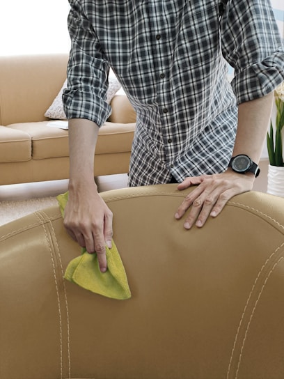 A person is seen cleaning a beige leather sofa with a yellow cloth. The individual is wearing a plaid shirt and a watch. The background includes a beige couch with cushions and a plant, indicating a living room setting.