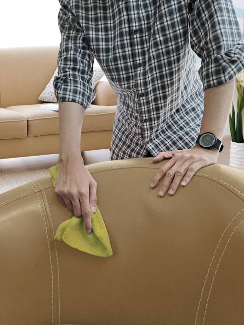 A person is seen cleaning a beige leather sofa with a yellow cloth. The individual is wearing a plaid shirt and a watch. The background includes a beige couch with cushions and a plant, indicating a living room setting.
