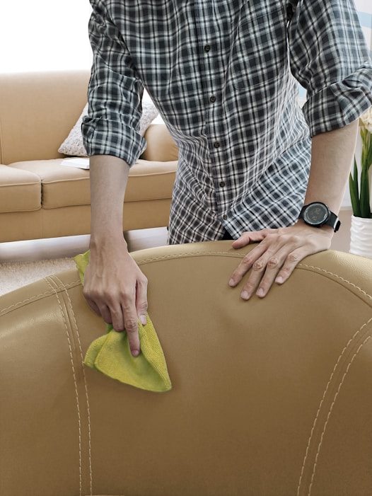 A person is seen cleaning a beige leather sofa with a yellow cloth. The individual is wearing a plaid shirt and a watch. The background includes a beige couch with cushions and a plant, indicating a living room setting.