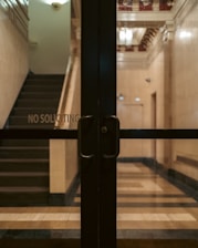 Glass doors with handles are situated in front of a dimly lit lobby area with visible stairs and a 'No Soliciting' sign. The interior features a tiled floor and decorative moldings on the ceiling.