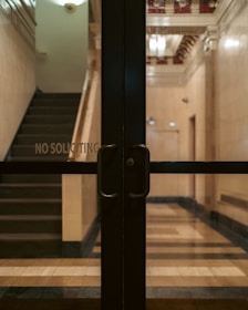 Glass doors with handles are situated in front of a dimly lit lobby area with visible stairs and a 'No Soliciting' sign. The interior features a tiled floor and decorative moldings on the ceiling.