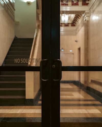 Glass doors with handles are situated in front of a dimly lit lobby area with visible stairs and a 'No Soliciting' sign. The interior features a tiled floor and decorative moldings on the ceiling.