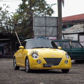 A bright yellow compact car features distinctive eyelash decorations on its headlights, adding a playful and quirky touch. The car is parked on a rough, unpaved surface, with a backdrop of urban elements including a green vehicle and assorted signage. Trees and a utility pole are visible in the background, suggesting an outdoor, possibly public or commercial setting.