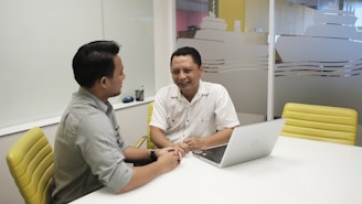 Two people are seated at a white table in an office setting, engaging in a conversation. One person is wearing a gray shirt, and the other is in a white shirt. A laptop is open on the table in front of them, and there are yellow chairs around the table. The background shows a frosted glass wall with designs resembling ships.