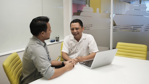 Two people are seated at a white table in an office setting, engaging in a conversation. One person is wearing a gray shirt, and the other is in a white shirt. A laptop is open on the table in front of them, and there are yellow chairs around the table. The background shows a frosted glass wall with designs resembling ships.