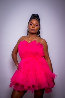 A person stands confidently wearing a vibrant pink, layered dress. The dress is made of a sheer, flowing fabric. The individual has braided hair styled in an updo and wears large hoop earrings. The makeup is bold, with noticeable eye shadow.