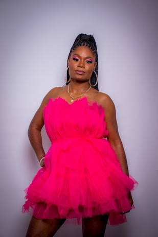 A person stands confidently wearing a vibrant pink, layered dress. The dress is made of a sheer, flowing fabric. The individual has braided hair styled in an updo and wears large hoop earrings. The makeup is bold, with noticeable eye shadow.