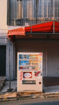 A vending machine is positioned on a sidewalk in front of a closed shop with a red awning. The machine displays a variety of bottled and canned drinks. Above the vending machine is a metal railing, and the image captures the setting during daylight with long shadows.