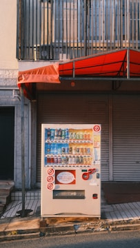 A vending machine is positioned on a sidewalk in front of a closed shop with a red awning. The machine displays a variety of bottled and canned drinks. Above the vending machine is a metal railing, and the image captures the setting during daylight with long shadows.
