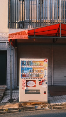A vending machine is positioned on a sidewalk in front of a closed shop with a red awning. The machine displays a variety of bottled and canned drinks. Above the vending machine is a metal railing, and the image captures the setting during daylight with long shadows.