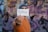 A person wearing an orange sweater and a gray beanie holds a card with the word 'Python' written on it. The background features blurred graffiti on a wall.