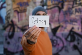 A person wearing an orange sweater and a gray beanie holds a card with the word 'Python' written on it. The background features blurred graffiti on a wall.