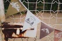 A row of triangular pennant flags is hanging on a metal fence. The flags have various outdoor-themed prints with phrases such as 