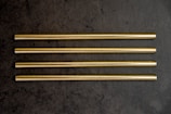 Four gold-colored metal straws are arranged in parallel on a dark grey textured surface.