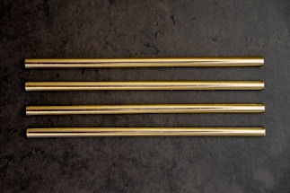 Four gold-colored metal straws are arranged in parallel on a dark grey textured surface.