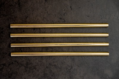 Four gold-colored metal straws are arranged in parallel on a dark grey textured surface.