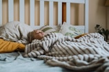 A person is sleeping under a thick, striped blanket on a bed. The bed is covered with several pillows, some with patterned pillowcases, set against a white headboard. Soft lighting and a plant in the background create a cozy atmosphere.