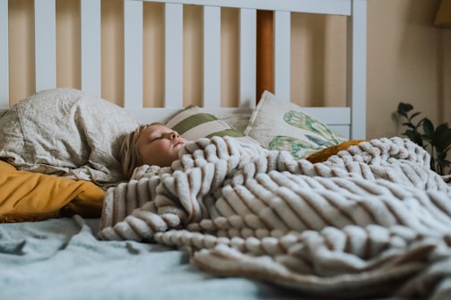 A person is sleeping under a thick, striped blanket on a bed. The bed is covered with several pillows, some with patterned pillowcases, set against a white headboard. Soft lighting and a plant in the background create a cozy atmosphere.