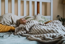 A person is sleeping under a thick, striped blanket on a bed. The bed is covered with several pillows, some with patterned pillowcases, set against a white headboard. Soft lighting and a plant in the background create a cozy atmosphere.