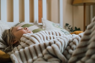 A child with light hair is peacefully sleeping, wrapped in a soft, striped blanket. The setting includes a bed with several pillows, and a warm, cozy atmosphere is created by soft, ambient lighting. A hint of green from some plants peeks out in the background.