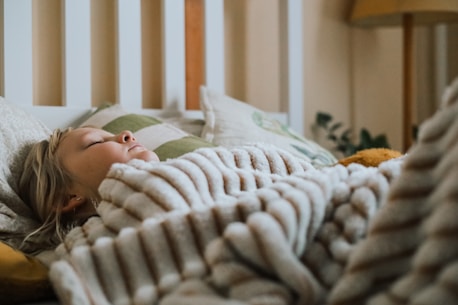 A child with light hair is peacefully sleeping, wrapped in a soft, striped blanket. The setting includes a bed with several pillows, and a warm, cozy atmosphere is created by soft, ambient lighting. A hint of green from some plants peeks out in the background.