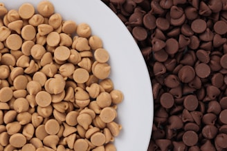 A plate is filled with a pile of light brown peanut butter chips, and a larger amount of dark brown chocolate chips surrounds it. The chips have a smooth, glossy texture.